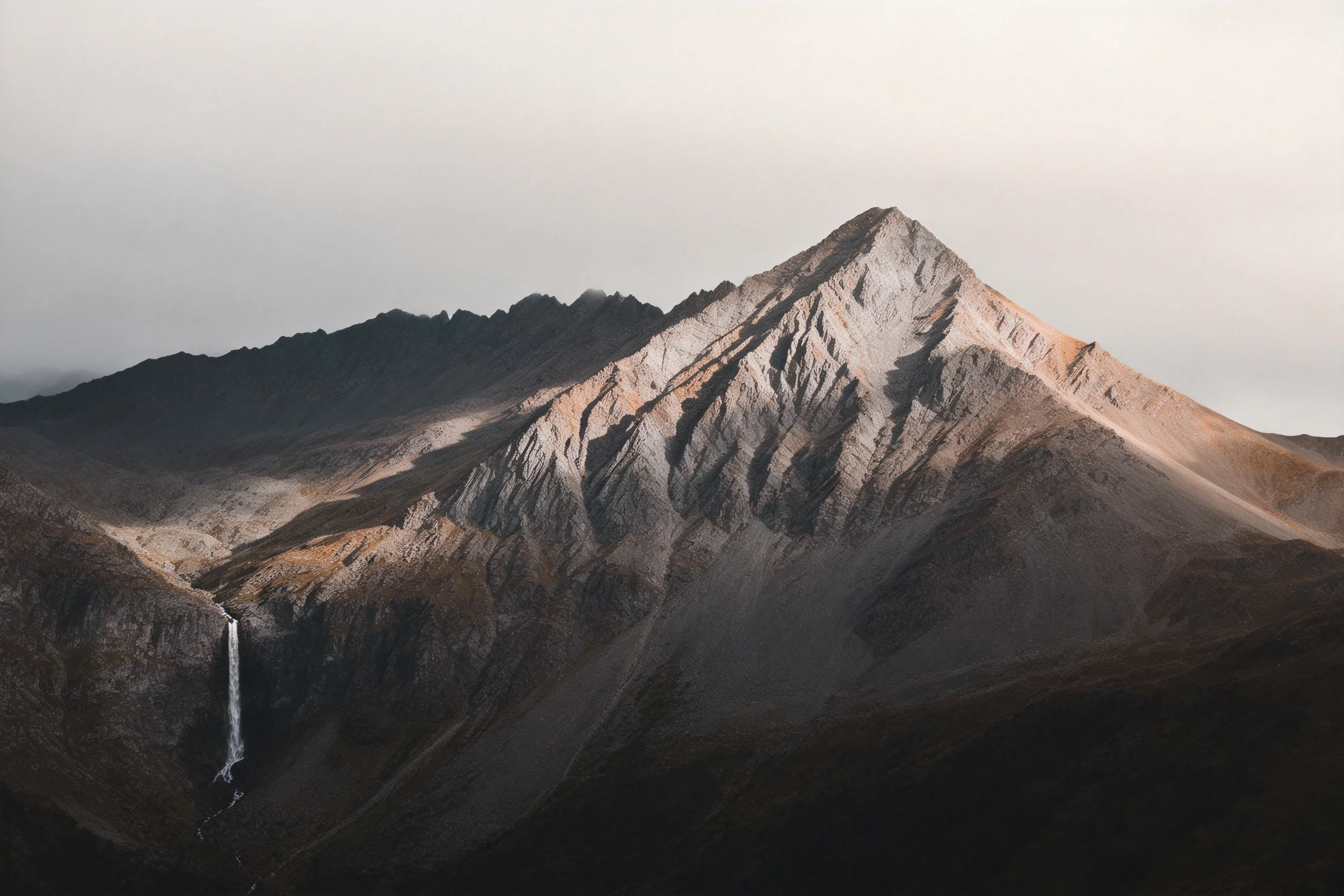 陡峭山地景观,岩石表面呈现出深浅不一的灰褐色纹理,主峰岩壁因光影形成明显明暗对比,瀑布水流垂直落下。