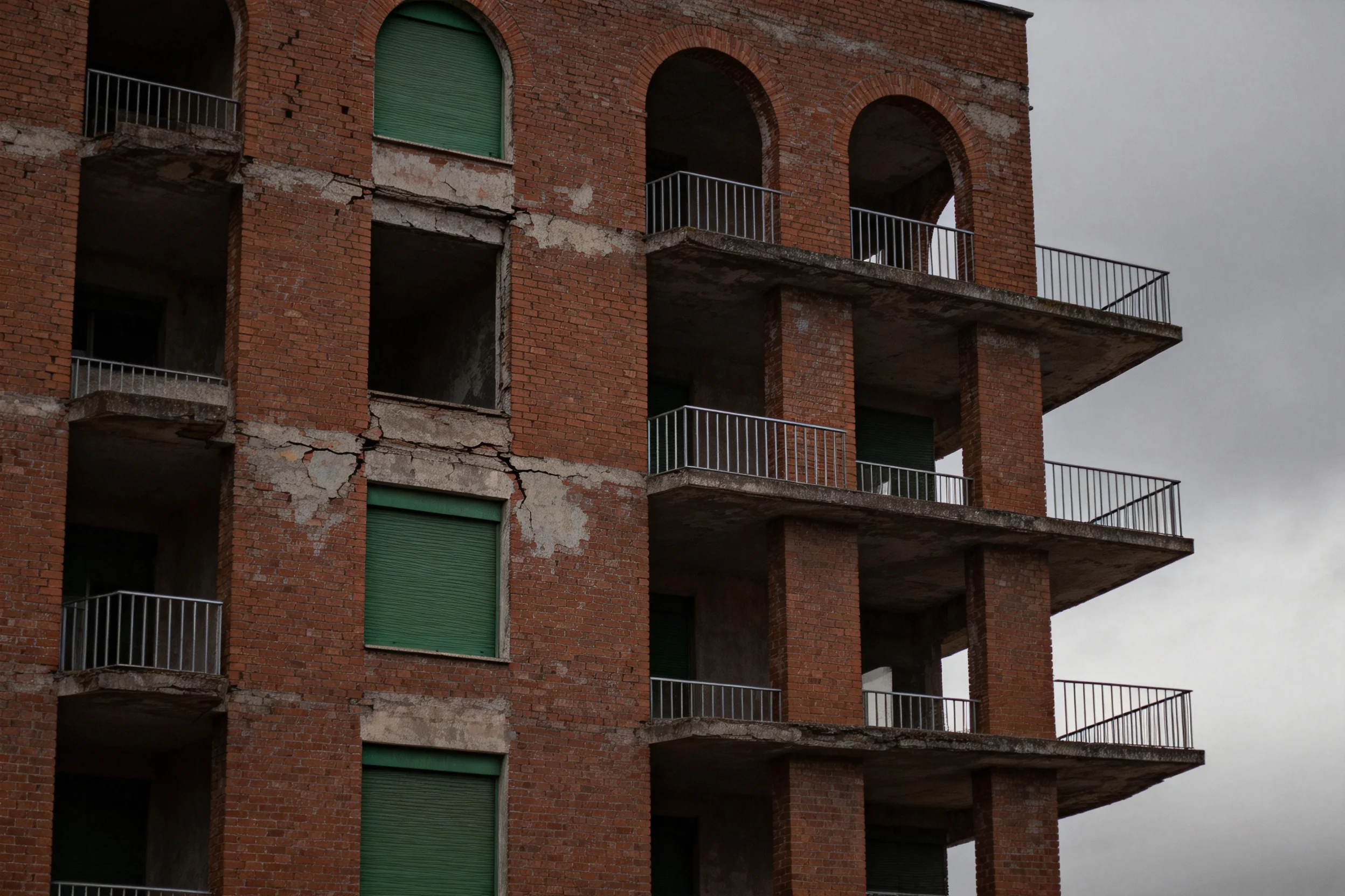 废弃红砖多层建筑外墙上布满裂缝,绿色卷帘门紧闭,显示这是一栋已废弃的建筑。