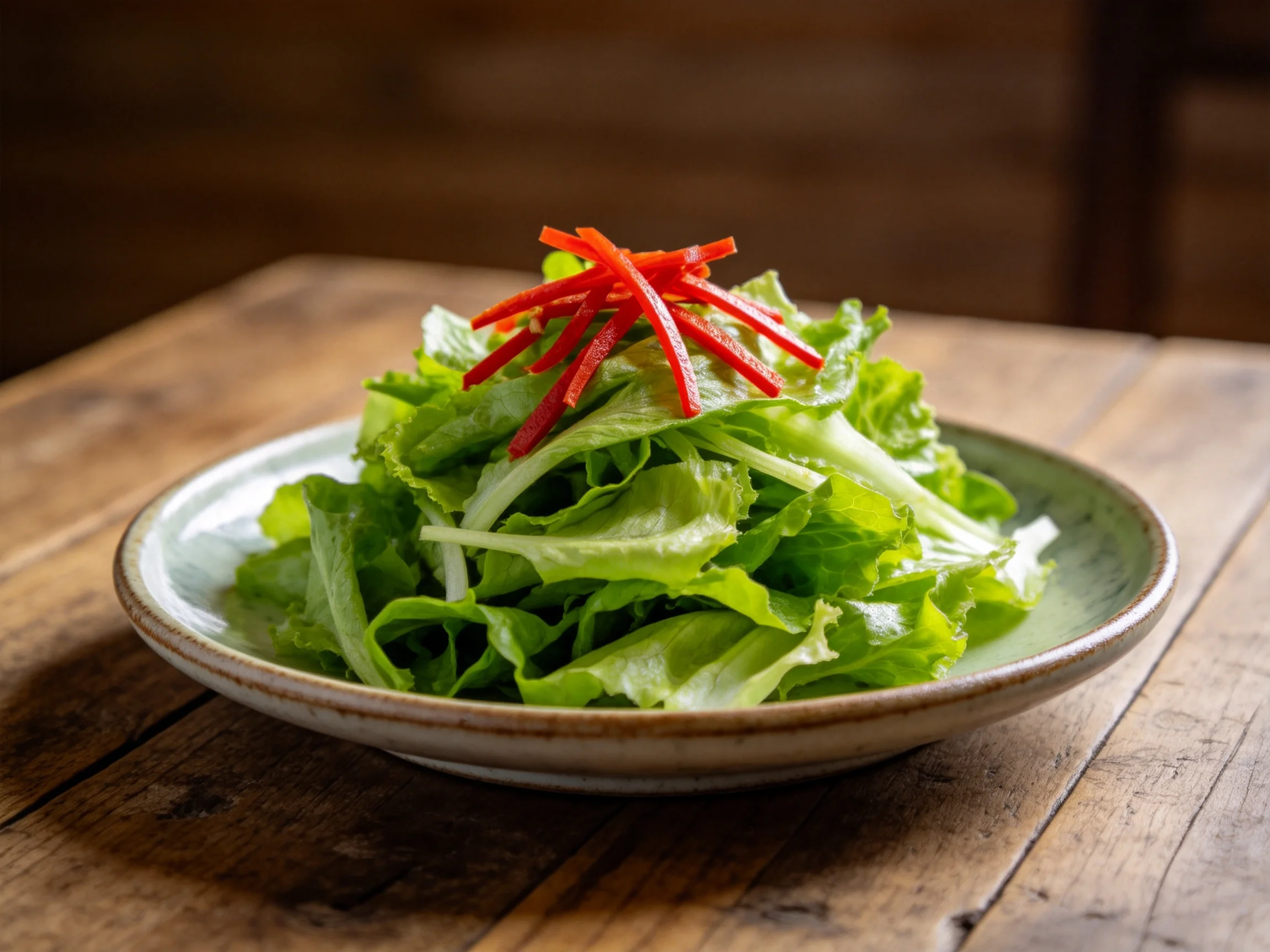 木桌上陶瓷盘里的凉拌生菜,翠绿生菜搭配红色辣椒丝,叶片饱满鲜嫩。