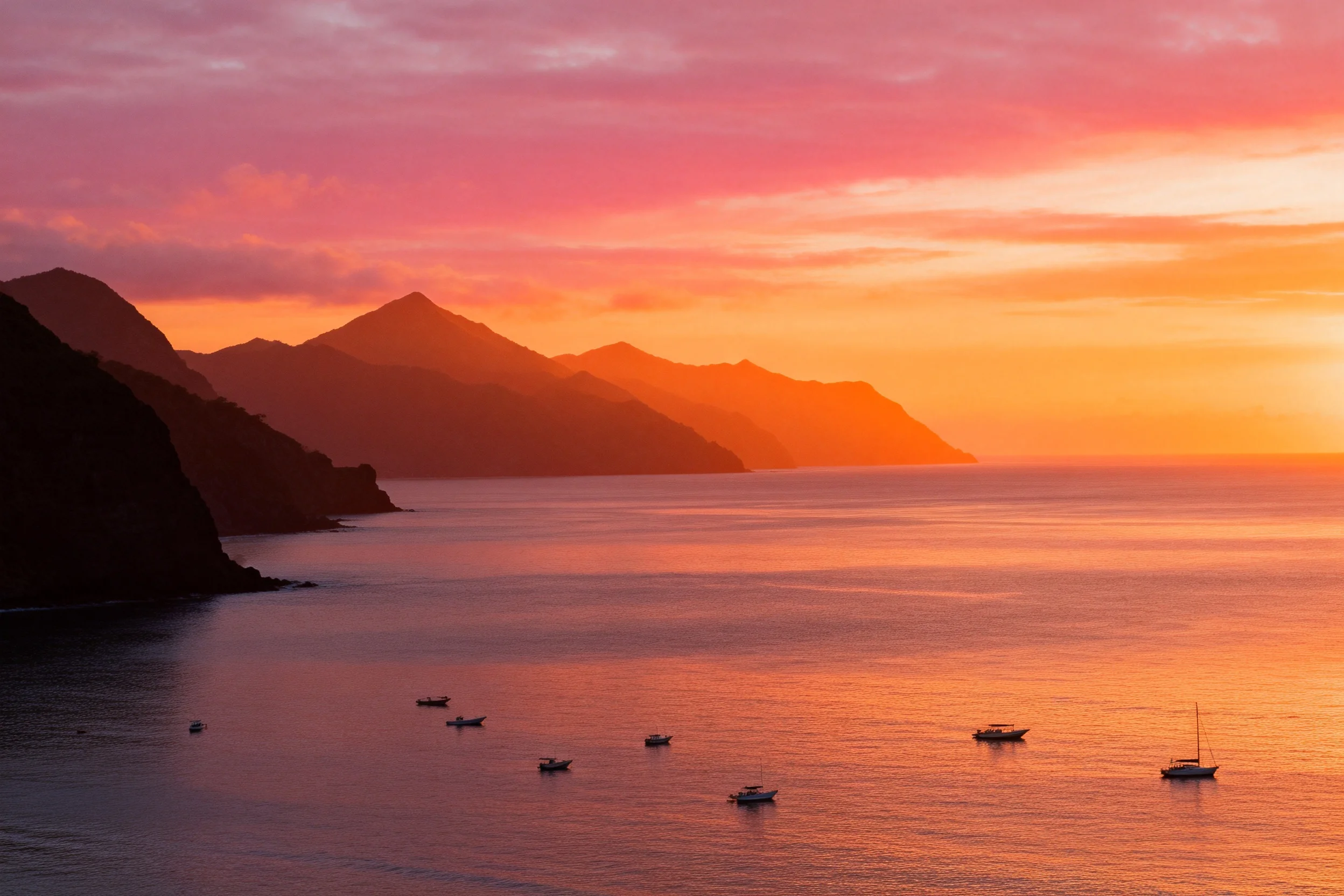 沿海山崖日落海岸风光里,多艘船只静静漂浮在泛着暖橙色波纹的海面上,远处山峦在晚霞映照下轮廓清晰。