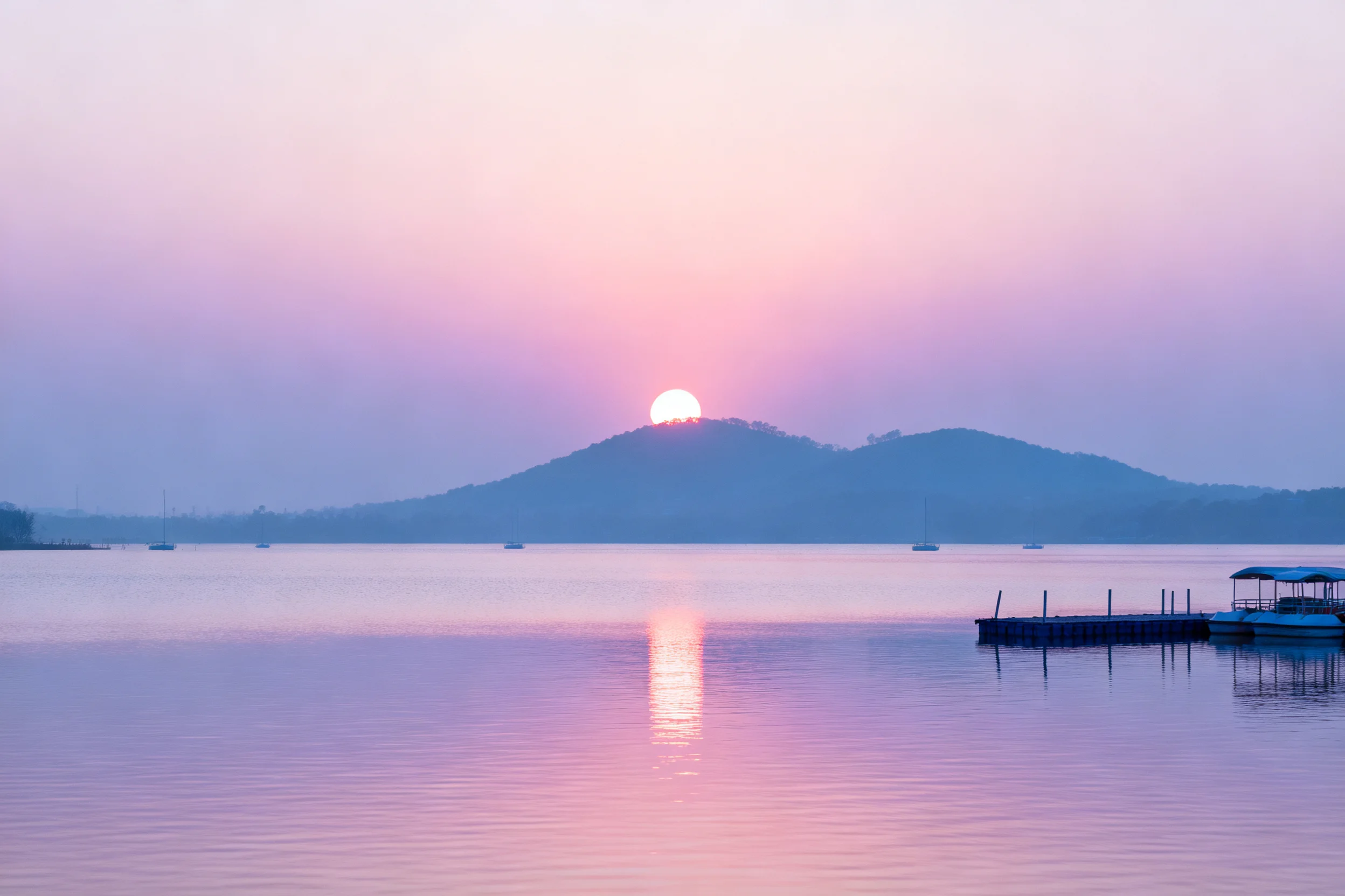 山水湖景中,太阳位于山巅位置,粉红色天空映照下,平静湖面有清晰的太阳倒影。